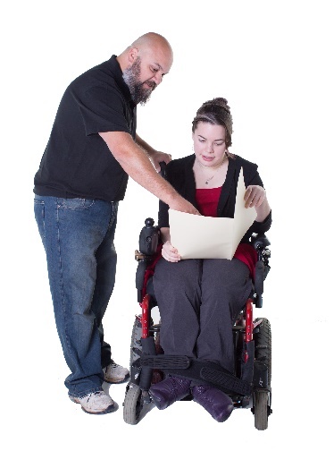 A person in a wheelchair holding a document. There is a person standing nearby, pointing to the document. 